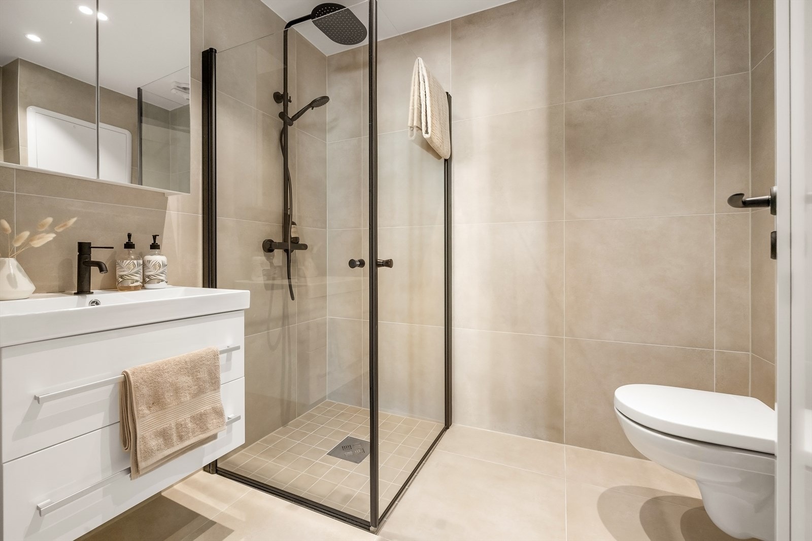 Lekkert og romslig bad som inneholder vegghengt toalett, helstøpt servant, skuffer under servant med slette fronter, dusjdører i klart glass og opplegg for vaskemaskin Galleribilde