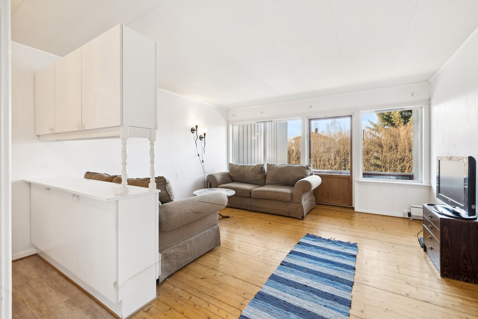 Stuen har lysmalte vegger og tregulv. Her er det tre store vinduer og en terrassedør som slipper dagslyset godt inn i rommet. Galleribilde