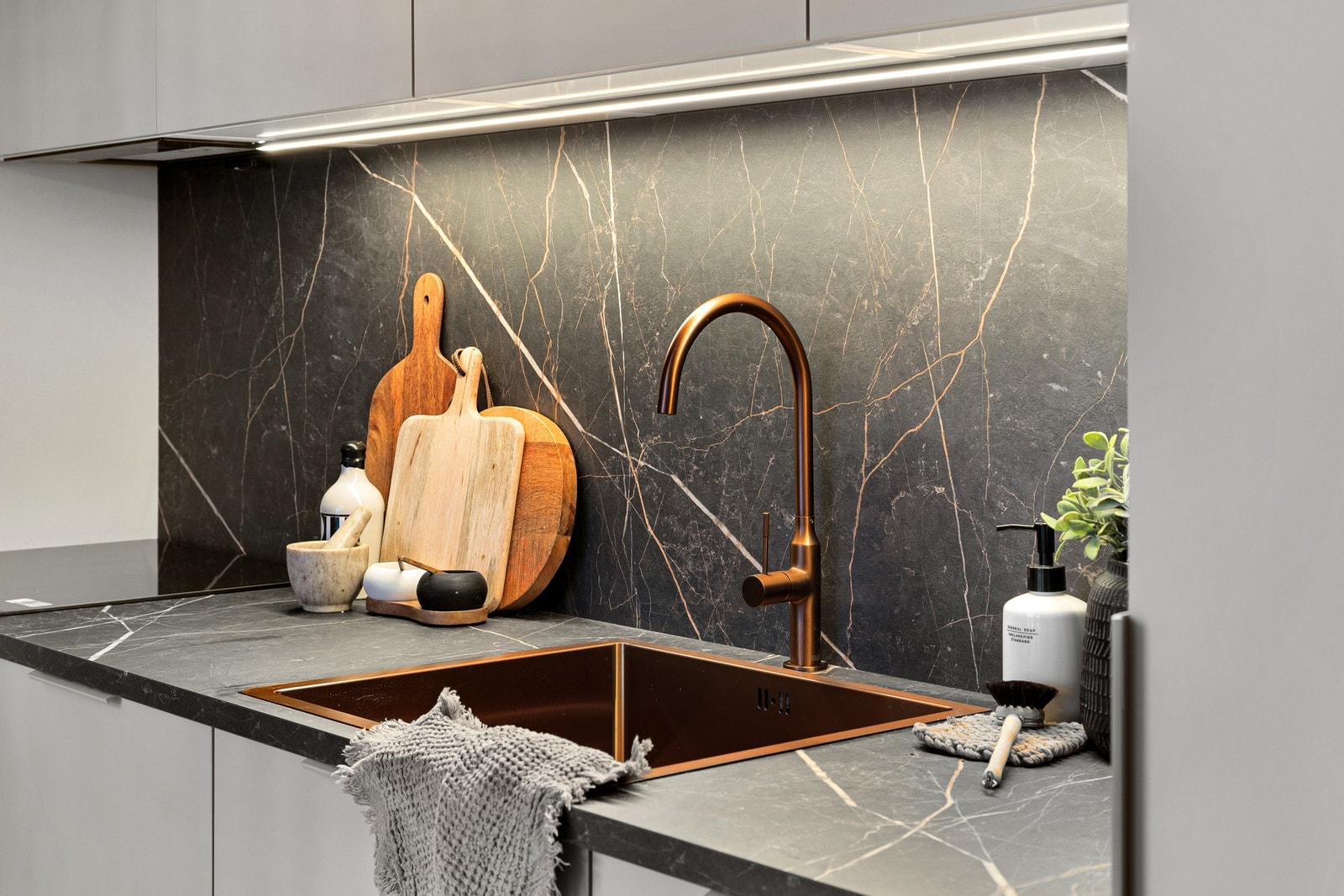 Kjøkkenet har mørk marmor laminat benkeplate med bronse vask og armatur, LED-stripe over benken og sprutplate i mørk marmor Galleribilde