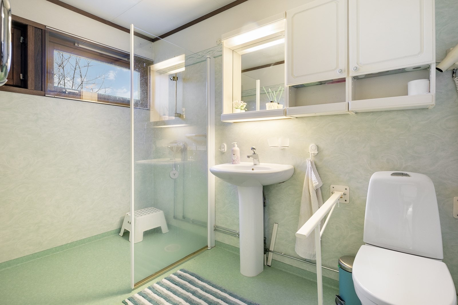 Bad med linoleum på gulv med varmekabler, servant, speil, overskap, og dusjhjørne med skillevegg i herdet glass Galleribilde