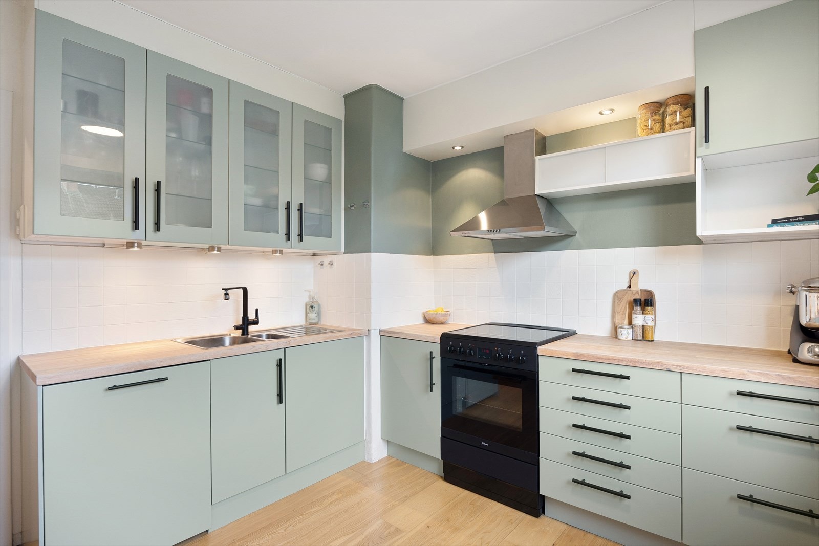 Kjøkkenet er romslig og har en moderne finish i en mintgrønn farge, kombinert med stilige firkantede fliser som backsplash. Innfelte downlights over kokeområdet og under vitrineskapene gir ekstra belysning. Galleribilde