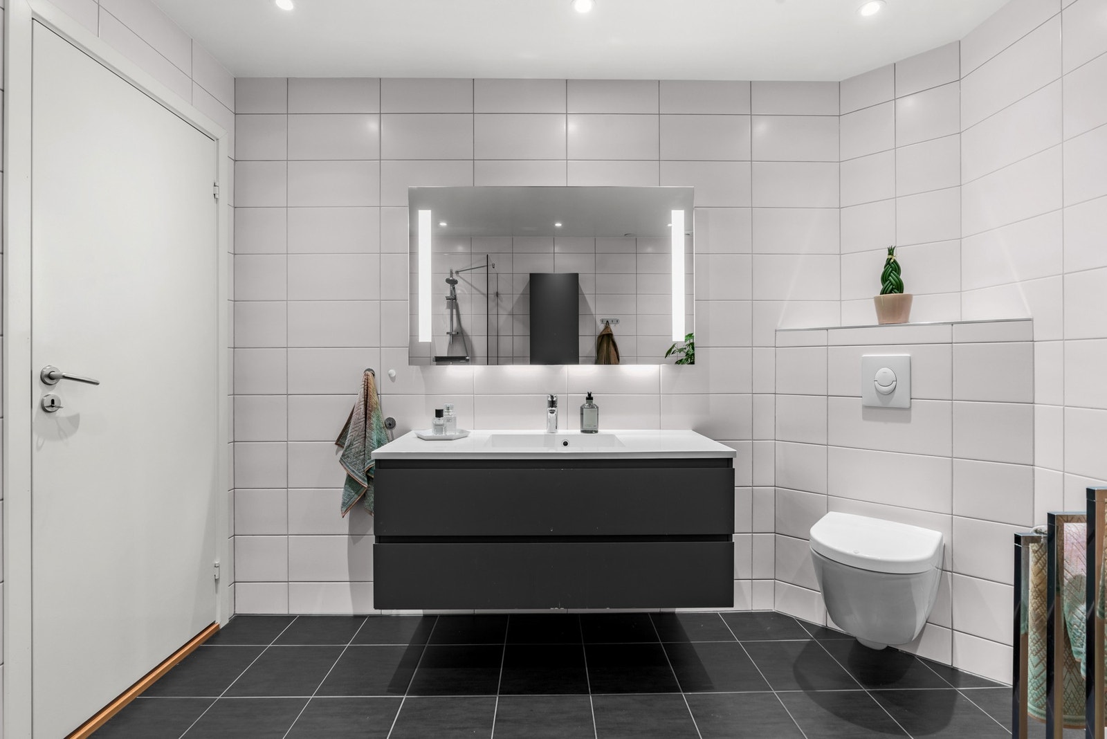 Det er utstyrt med heldekkende servant over skap med brede skuffer, tilhørende høyskap, speil med integrert belysning og vegghengt toalett. Galleribilde