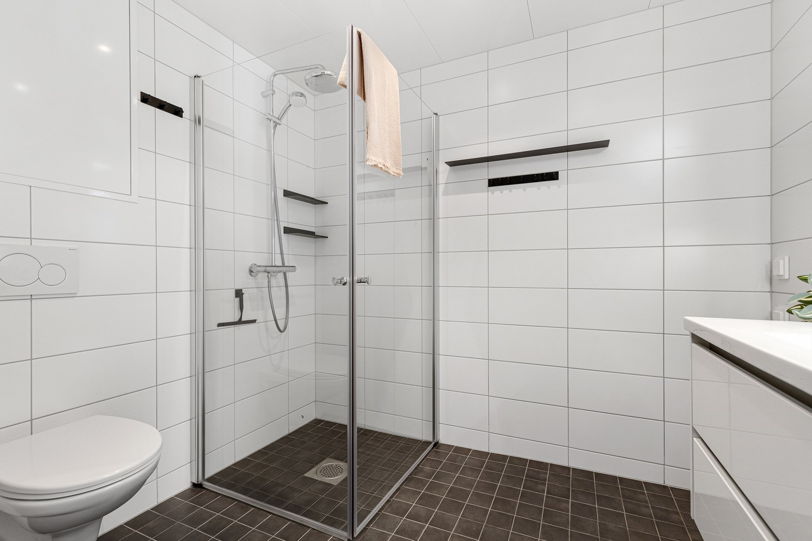 Videre er badet innredet med servantskap med overskap og speil med lys, vegghengt toalett og dusjhjørne med regndusj. Galleribilde