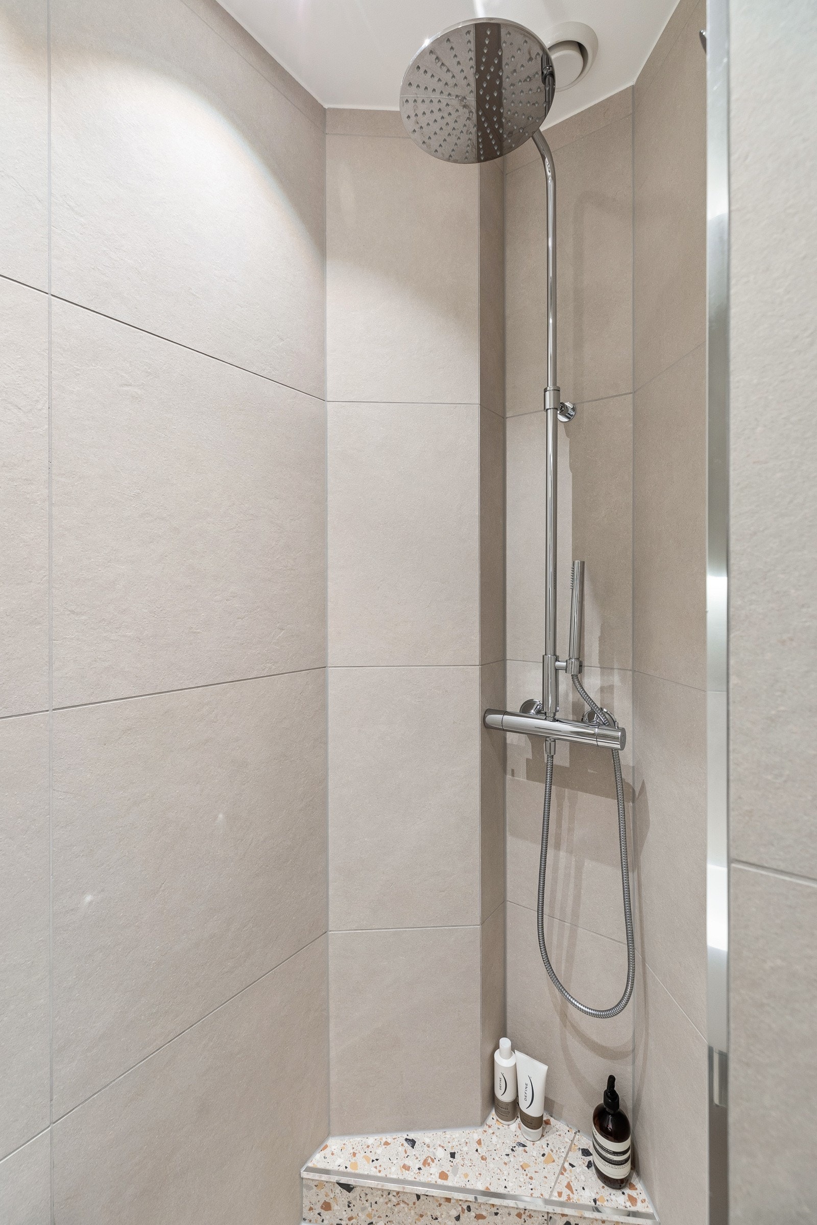 Baderomsinnredningen byr på servant med skapinnredning på vegg, speil med integrert belysning, åpent dusjhjørne med regnfallsdusj og toalett Galleribilde