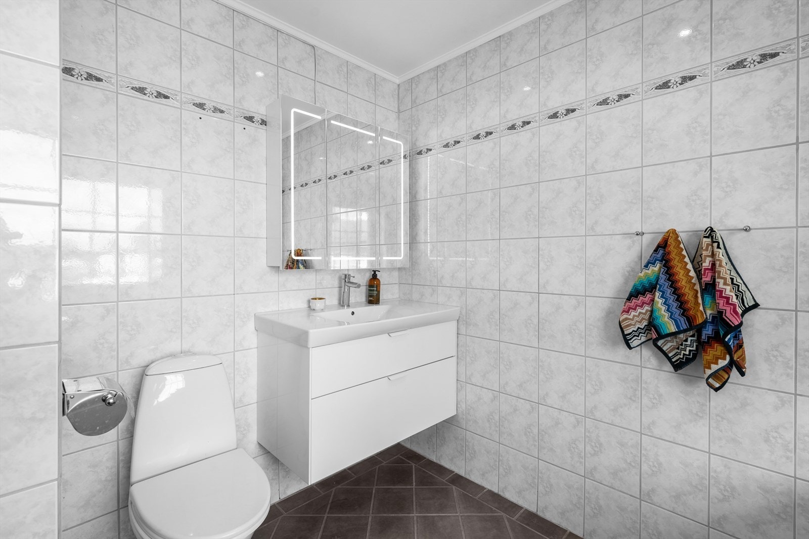 Baderomsinnredningen består av vegghengt skuffeseksjon med hvite, slette fronter, heldekkende servant og et stort speilskap med integrert belysning. Galleribilde