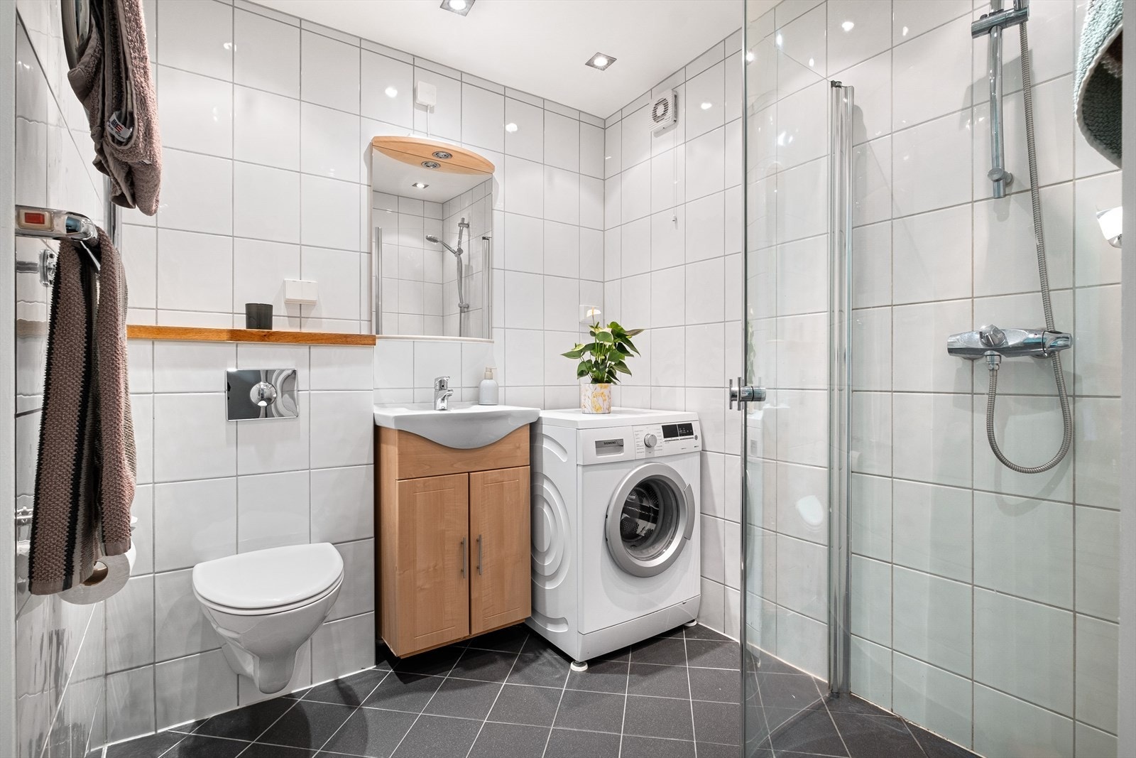 Pent flislagt bad med varmekabler i gulv. Badet er innredet med servant i innredning med skap, speil med belysning på vegg, dusj med innfellbare glassdører og vegghengt toalett Galleribilde