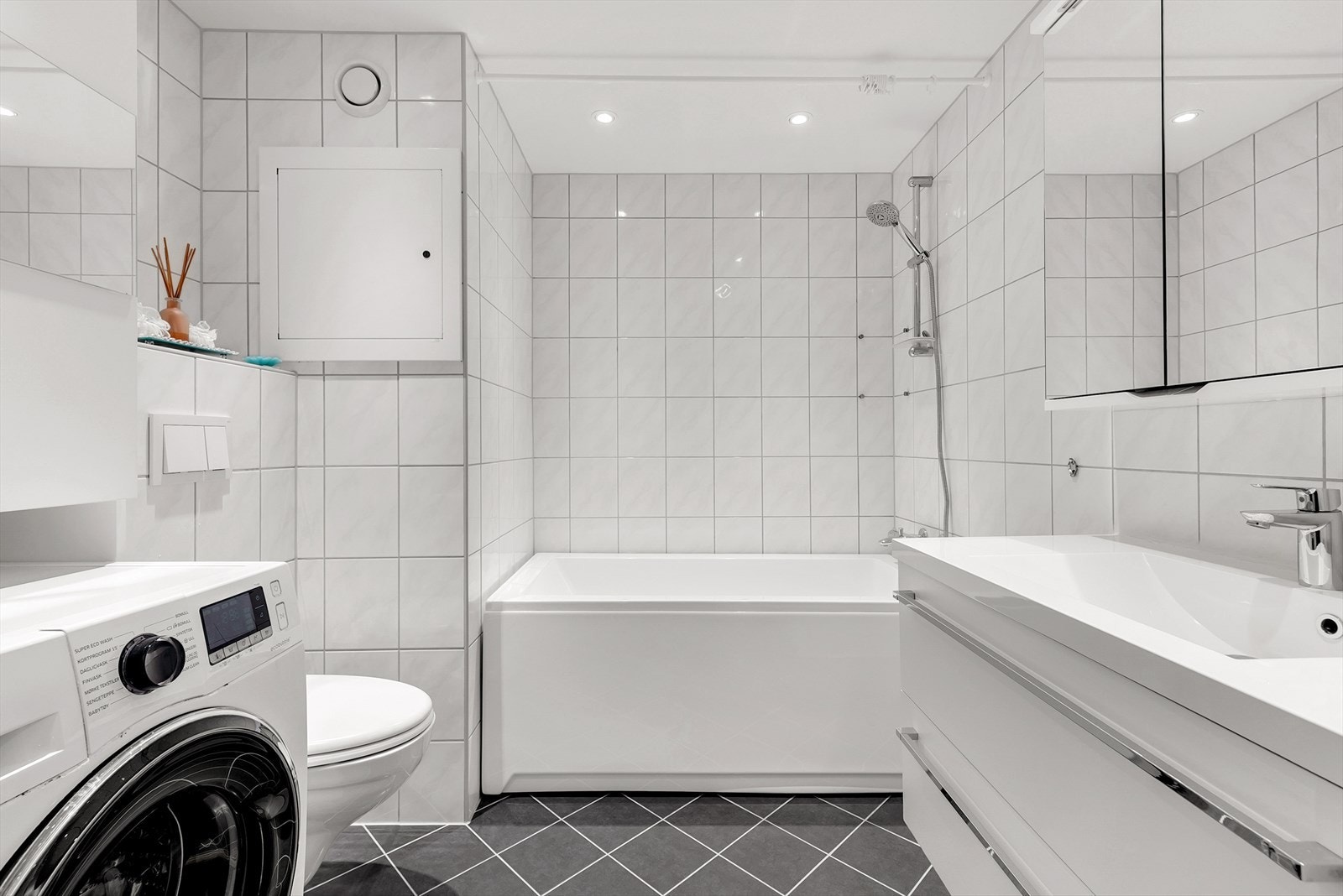 Flislagt bad som er innredet med badekar, vegghengt toalett, servant med skuffer og overskap med speil. Varmekabler i gulv. Galleribilde