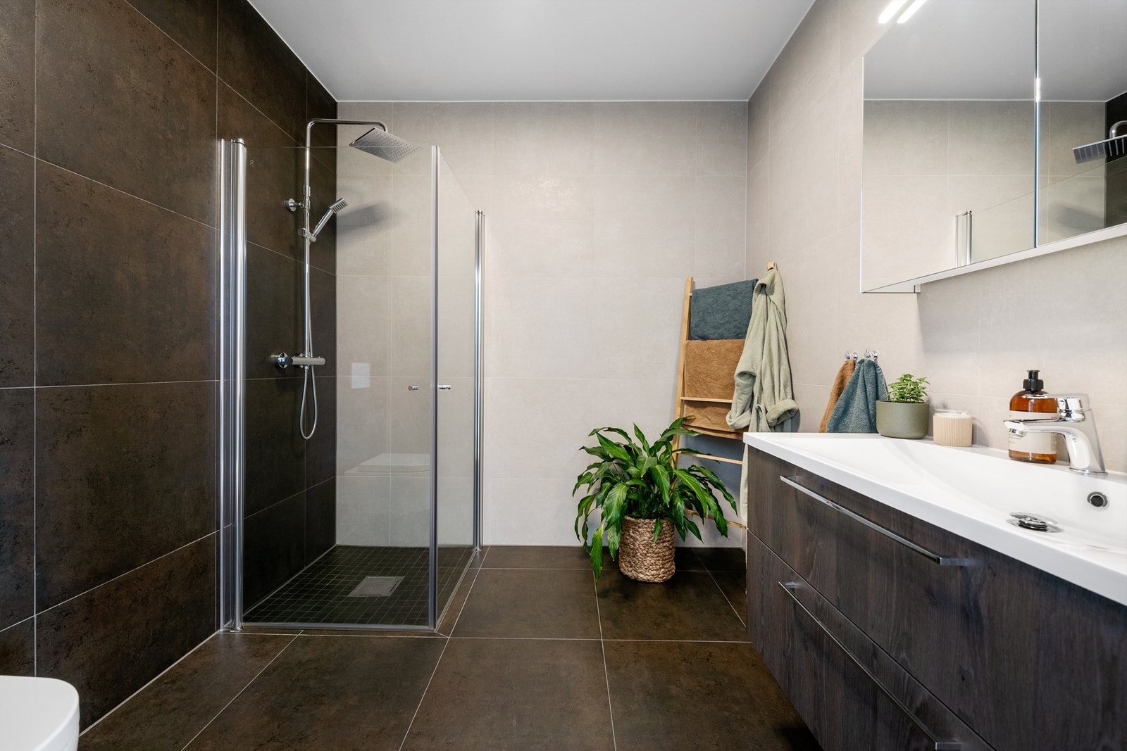 Flott flislagt bad med servantinnrending med skuffer, speilskap med dører over servant, dusjhjørne med glassdører og vegghengt dusjarmatur med hånddusj og regndusj Galleribilde