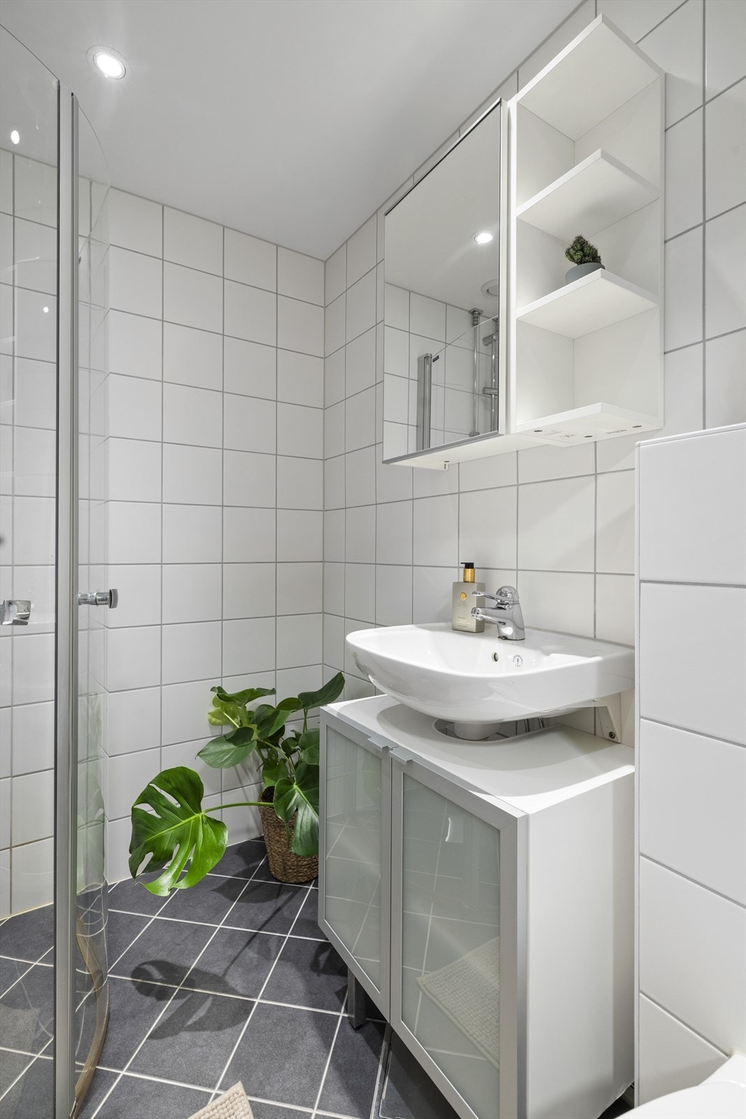 Badet er innredet med vegghengt toalett. og dusjhjørne med innfellbare glassdører. Baderomsinnredning med servant og overskap med speil Galleribilde