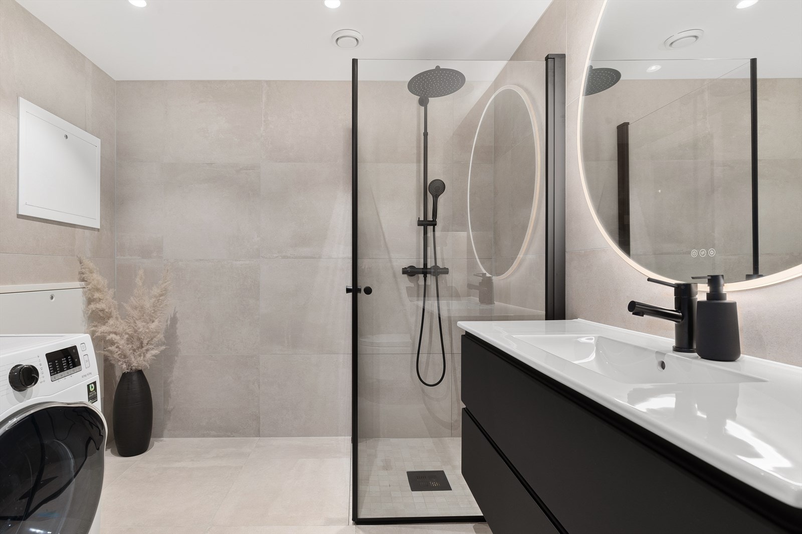 Badet er innredet med heldekkende servant nedfelt i skapinnredning med mørke slette fronter, speil med integrert ledlys, vegghengt toalett, dusjhjørne med dusjdører i glass samt opplegg og plass for vaskemaskin og tørketrommel Galleribilde
