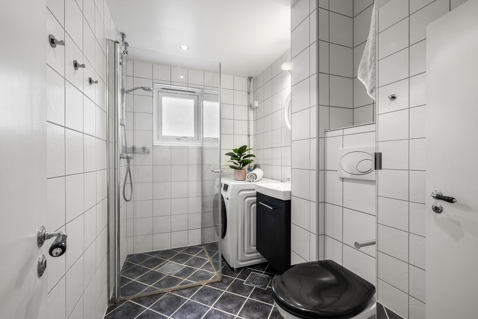 Badet har lyse veggfliser og mørkegulvfliser. Det er toalett, dusjhjørne, baderomsmøbel med speil og opplegg for vaskemaskin. Galleribilde