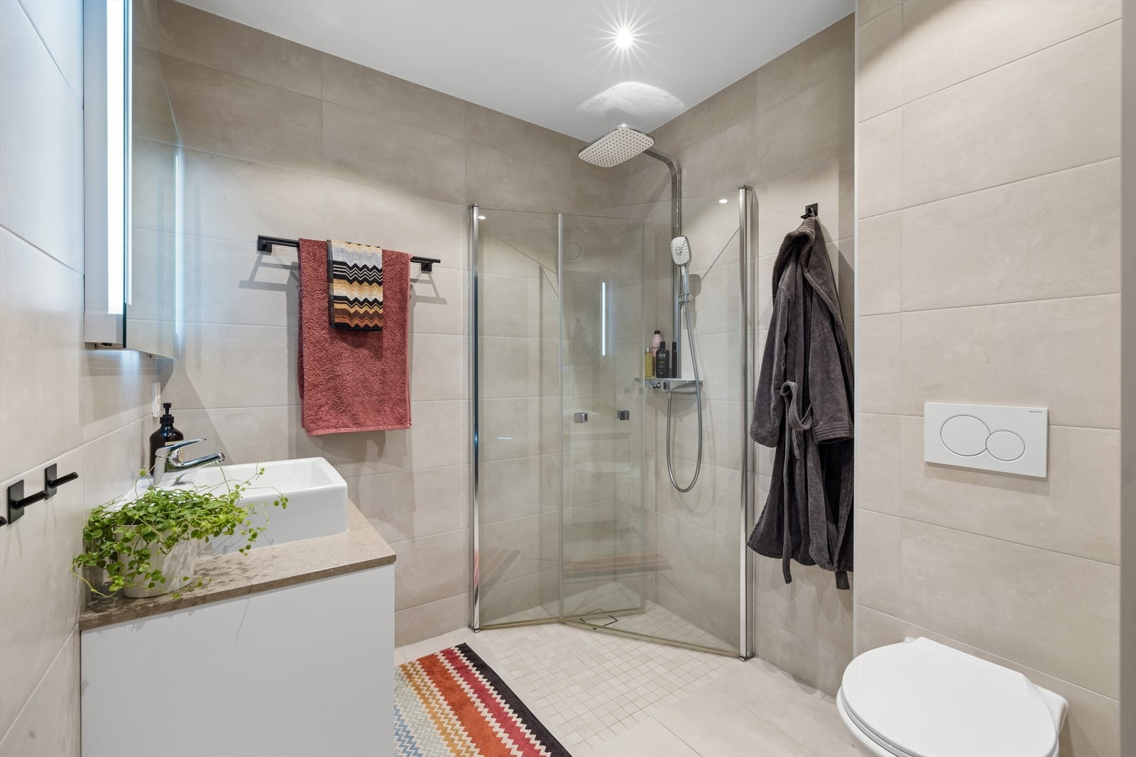 Stilrent og moderne bad som er flislagt med store fliser i lyse, duse farger. Badet er innredet med vegghengt wc, dusjsone med massasjedusj fra Oras Esteta wellfit med restitusjonsmodus og innfellbare glassdører. Galleribilde