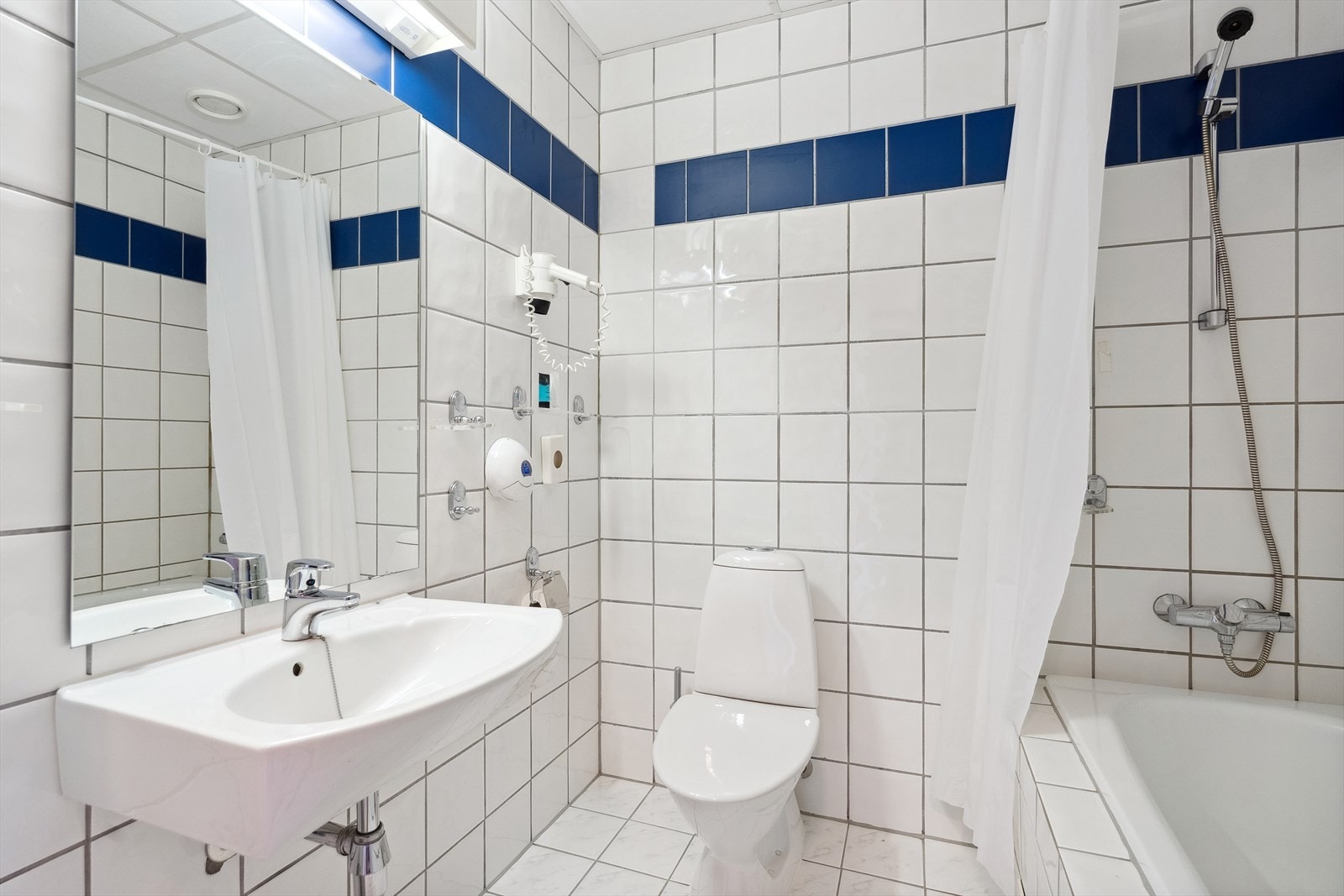 Bad med flisbelagte overflater, med varmekabler i gulvet. Det er montert vegghengt porselenservant, gulvstående toalett, vegghengt speil og badekar med dusjgarnityr på vegg. Galleribilde