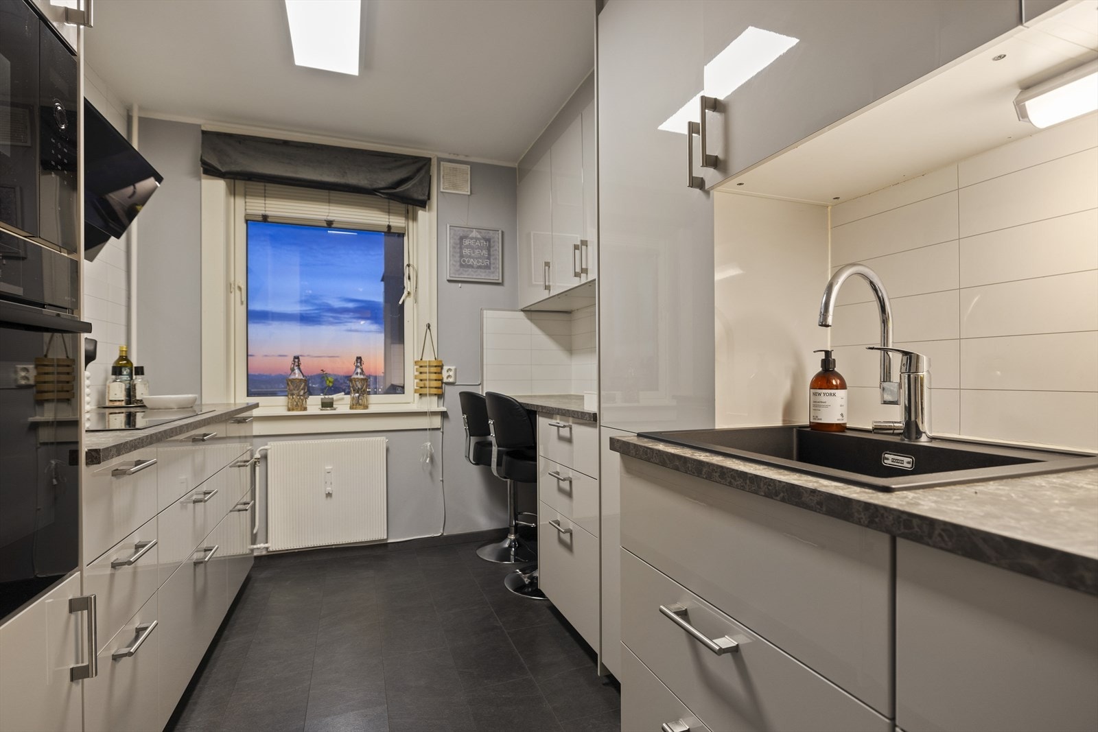 Glatte fronter i hvit høyglans, og laminat benkeplate med nedfelt kum i kompositt. Kjøkkenet er fullt utstyrt med integrerte hvitevarer fra Bosch, inkludert induksjonstopp, stekeovn, mikrobølgeovn og kjøleskap. Galleribilde