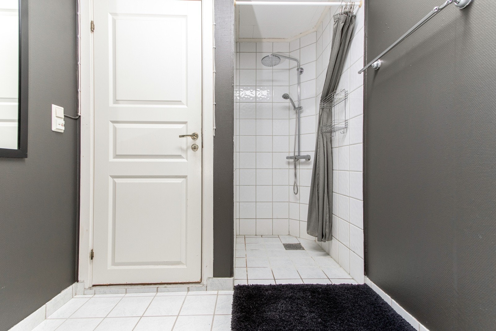 Gulvet har hvite fliser med varme, mens veggene er kledd med grå våtromstapet og matchende fliser. På badet er det toalett, opplegg for vaskemaskin og en åpen dusjnisje med regnfallsdusj. Galleribilde