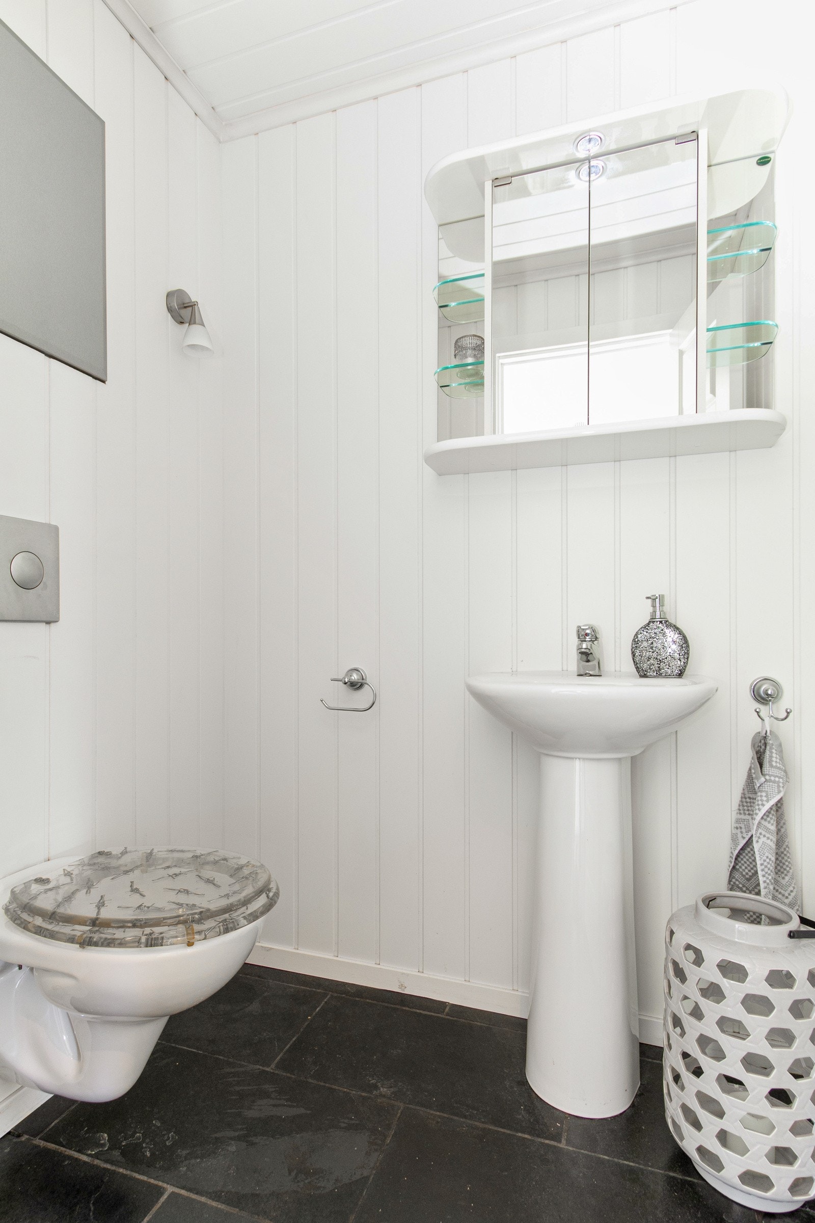 Wc-rommet har flislagt gulv, vegghengt toalett og en søyleservant med tilhørende speilskap. Galleribilde