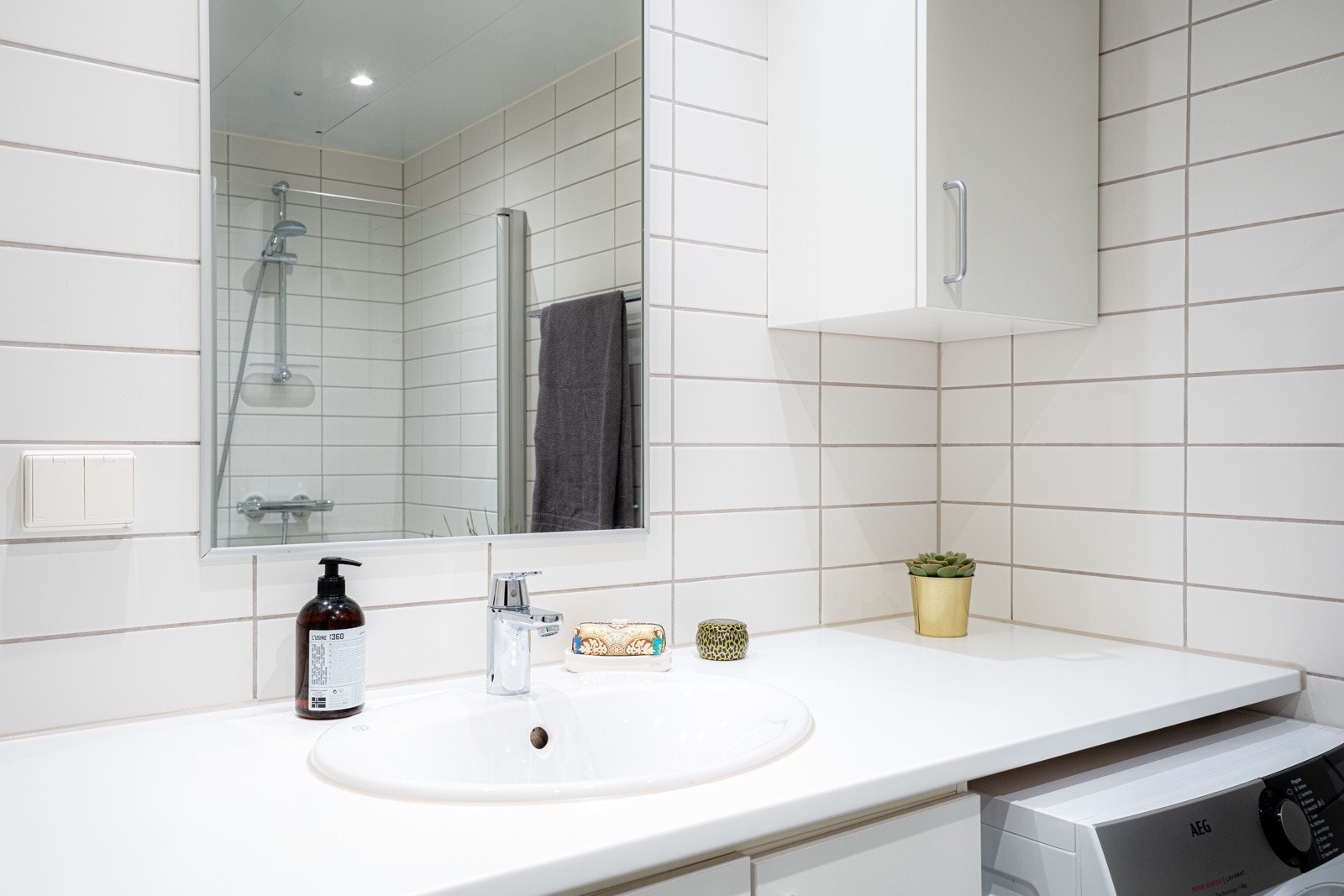 Badet er utstyrt med dusj med innfellbare glassvegger, servantseksjon med skap og heldekkende porselensservant, overskap, speil med lys og stikkontakter, vegghengt wc og opplegg for vaskemaskin og tørketrommel. Galleribilde