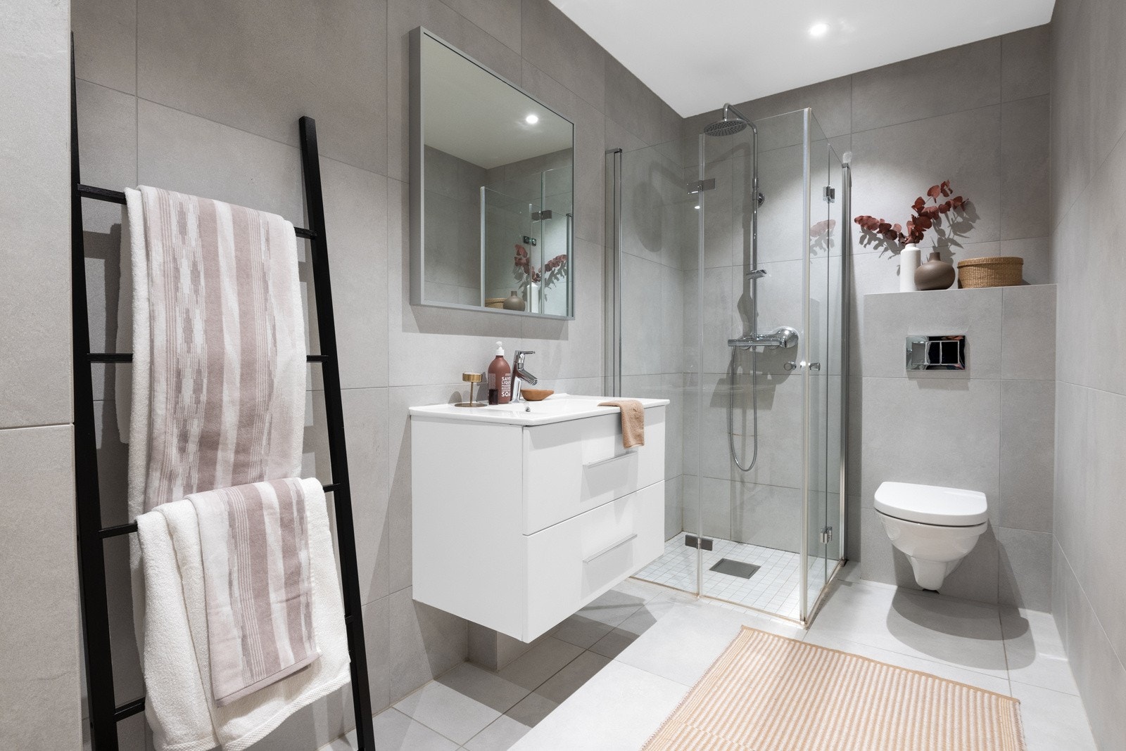 Lyst, delikat og flislagt baderom innredet med dusjnisje, servant med skuffeseksjon, klosett og opplegg for vaskemaskin og tørketrommel. Galleribilde