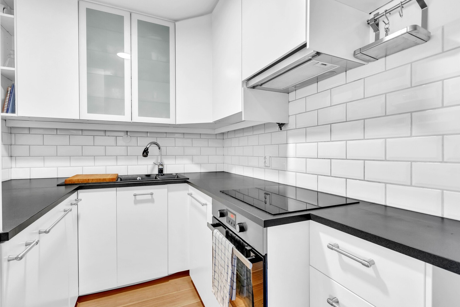 Her får du en delvis åpen kjøkken og stue løsning. Kjøkkeninnredning med hvite slette fronter, mørk benkeplate av laminat og hvite fliser på vegg mellom benkeplate og overskap. Galleribilde