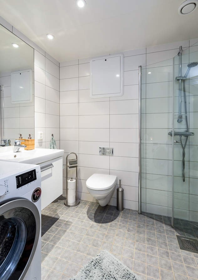 Kombinert bad og vaskerom med flis på golv og veggar. Badet er innreiia med servant i underskåp, spegel, vegghengt toalett, veggmontert dusj med dusjveggar og vaskemaskin. Galleribilde
