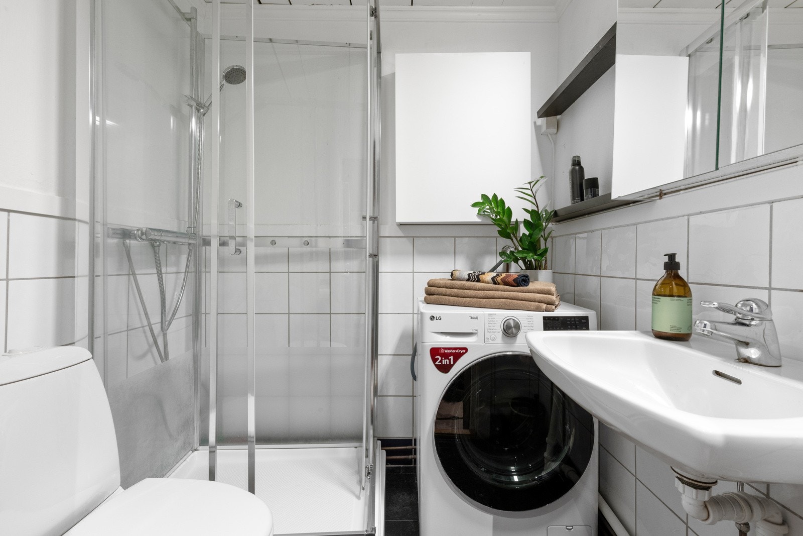 Lyst, flislagt baderom som inneholder dusjkabinett, speilskap, servant, toalett og opplegg for vaskemaskin. Galleribilde