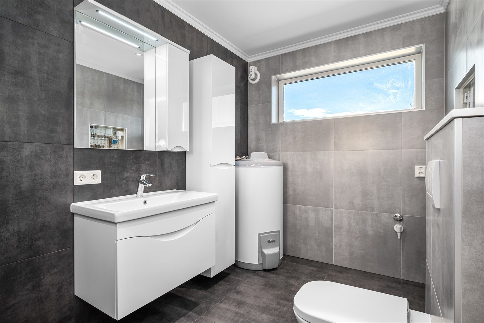 Badet er utstyrt med baderomsinnredning, veggmontert WC, varmtvannsbereder, opplegg for vaskemaskin og dusjhjørne Galleribilde