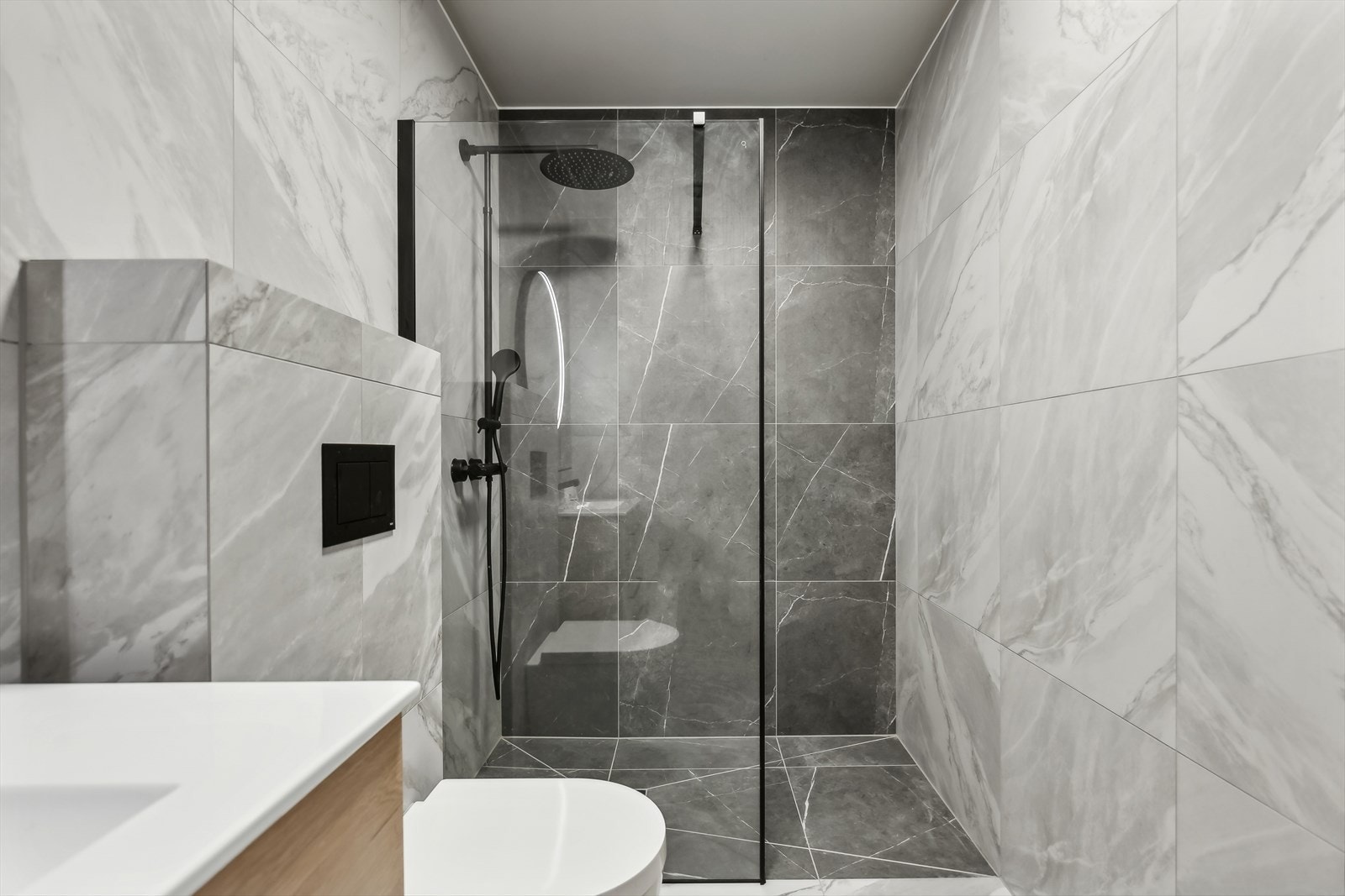 Badet inneholder vegghengt toalett, servantskap med oppbevaring, rundt speil og dusjnisje. Galleribilde