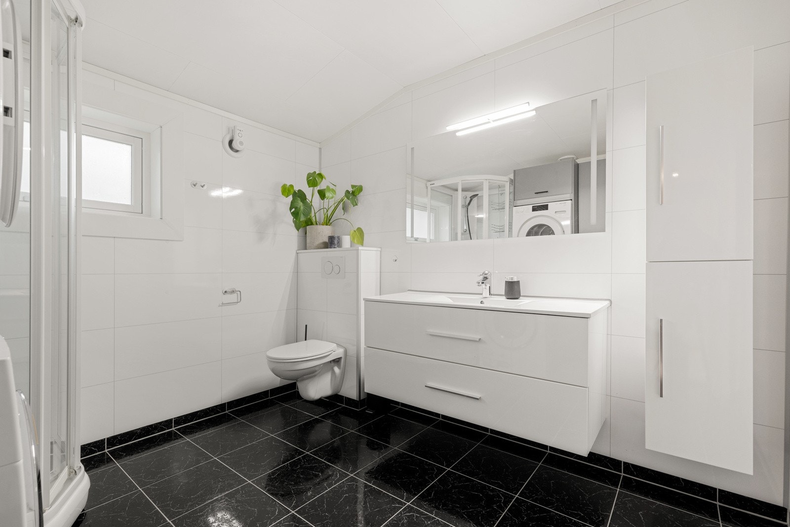 Delikat baderom som holder et tidløst preg og er bestående av servant med underskuffer, vegghengt speil med lys, dusjkabinett og vegghengt toalett Galleribilde