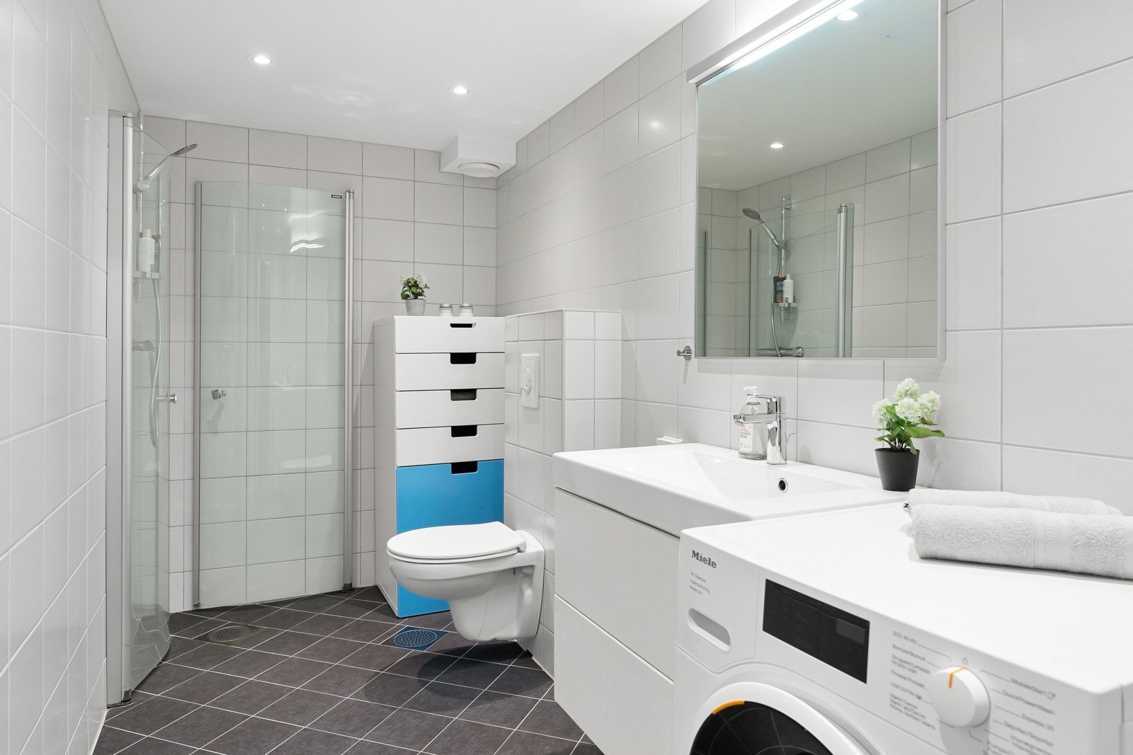 Fint, flislagt baderom som består av dusjhjørne, servant over innredning, speil og vegghengt toalett. Galleribilde