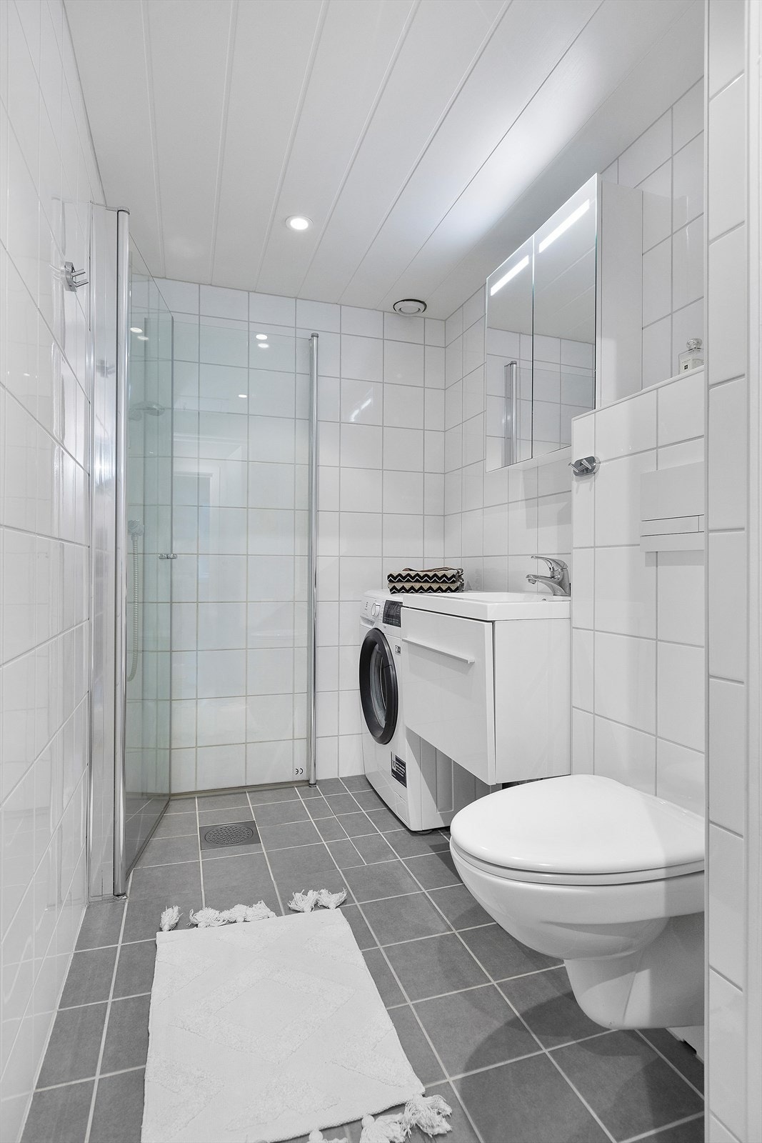 Badet har innredning med nedfelt servant, veggmontert toalett, dusjvegger/hjørne og opplegg for vaskemaskin. Fliser på gulv med varmekabler i gulvet. Downlights i taket. Galleribilde