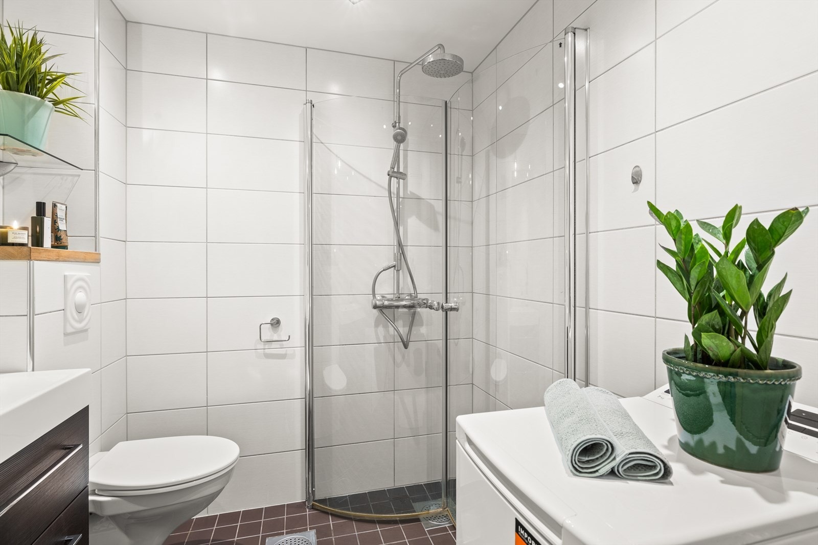 Badet har dusjhjørne med praktiske innfellbare dusjdører, vegghengt toalett, servant med oppbevaring og opplegg for vaskemaskin Galleribilde