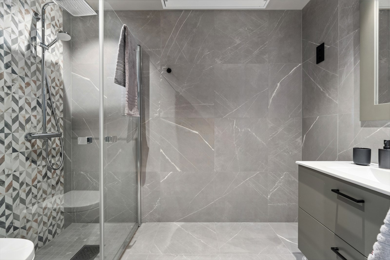 Delikat baderom utstyrt med servantskap med helstøpt servantplate, veggmontert klosett og dusjhjørne med glassdører. Galleribilde