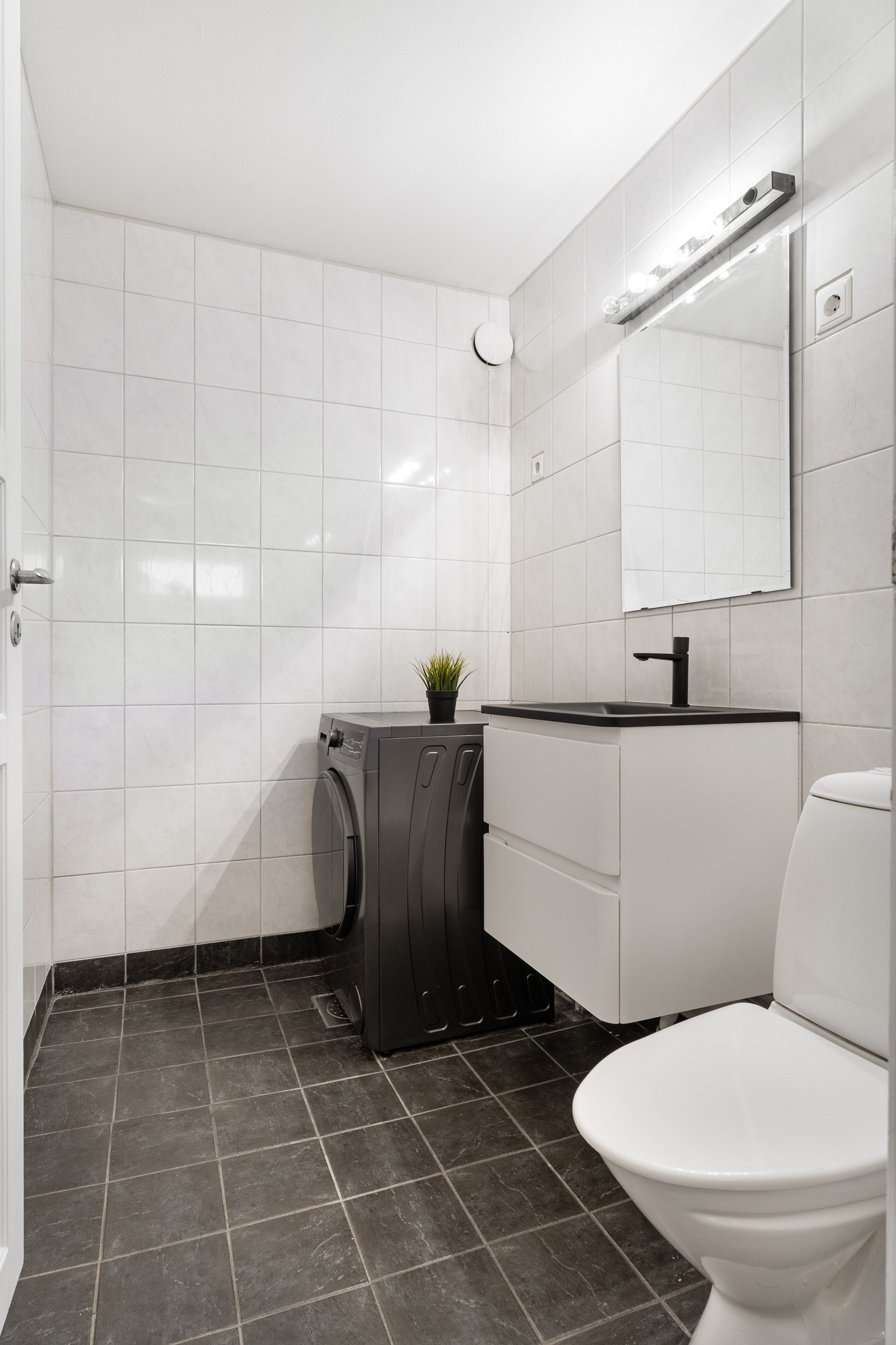 Videre fordeler entréen seg til et praktisk vaskerom, utstyrt med opplegg for vaskemaskin, en pen baderomsinnredning med skuffeseksjoner og toalett. Galleribilde