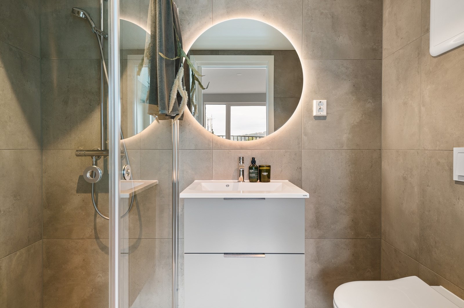 Lekkert bad med 60x30 fliser på gulv med gulvvarme og 60x60 fliser på vegg. Gulvet i dusjsonen har 5x5 fliser. Innredningen består av servant, grå skuffeseksjon og speil med integrert belysning, veggmontert toalett og dusjhjørne med regnfallsdusj. Galleribilde