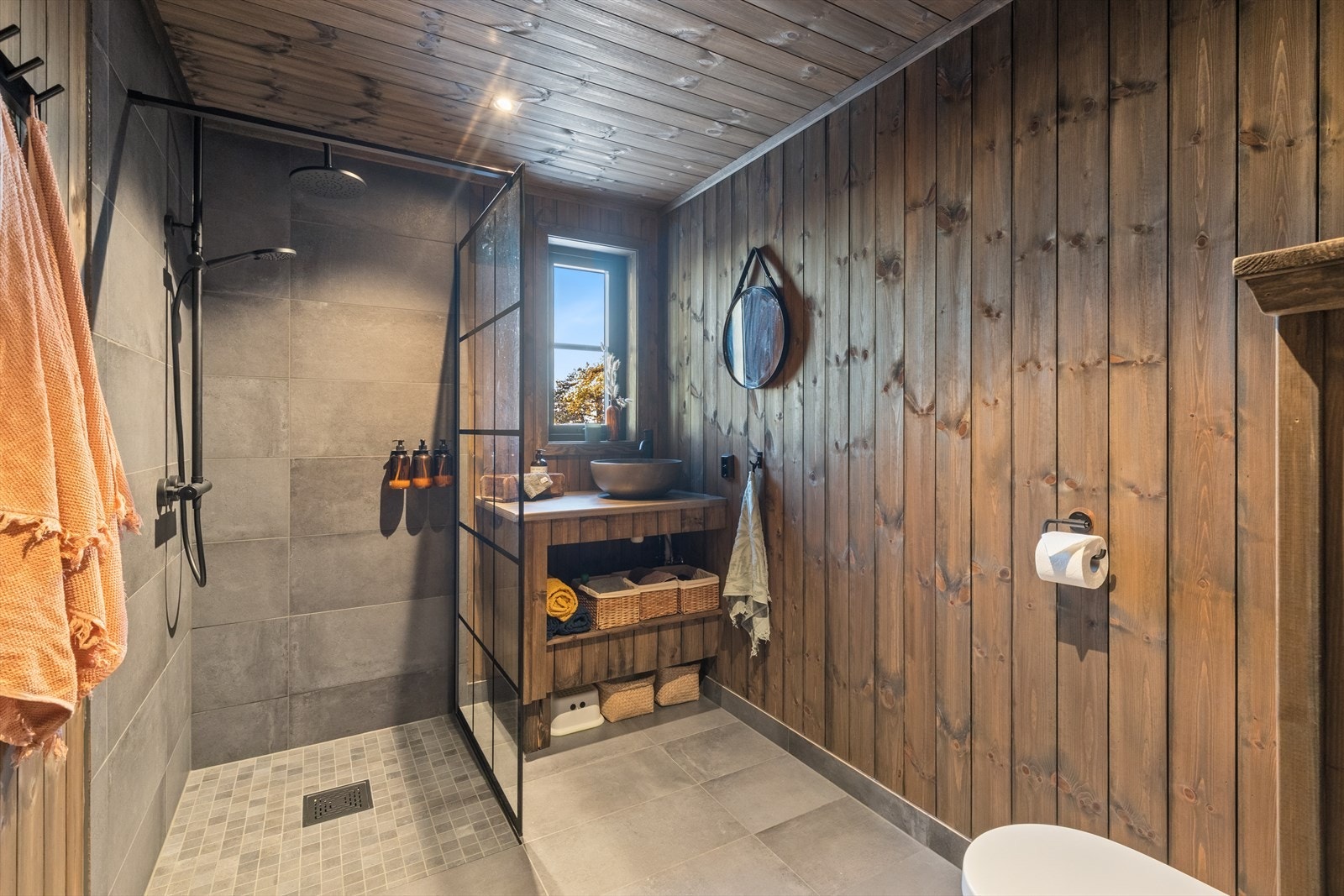 Badet har dusjhjørne med tak/regnfallsdusj og hånddusj, skillevegg mot servant i glass med flislagte overflater i dusjsone. Galleribilde