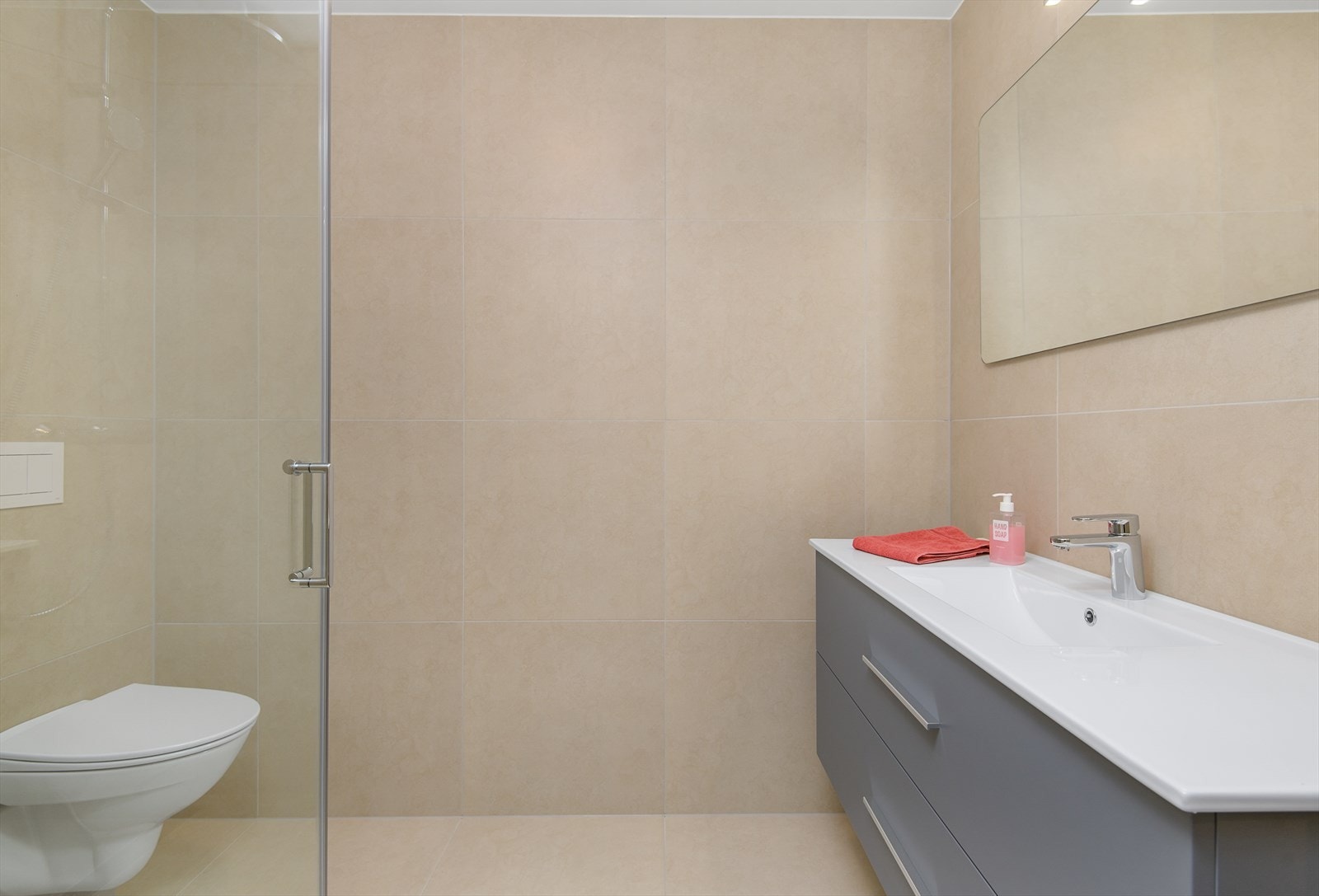 Hovedbad med storformat flis på gulv og vegg i fargen "Ivory". Innredning bestående av servant med underskuffer i grå utførelse og speil med lysarmatur. Galleribilde