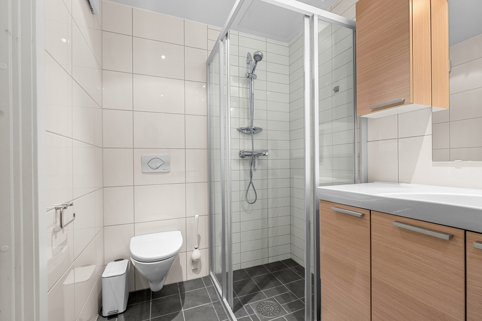 Badet er utstyrt med vegghengt toalett, dusjhjørne med dører og servantinnredning med speil Galleribilde