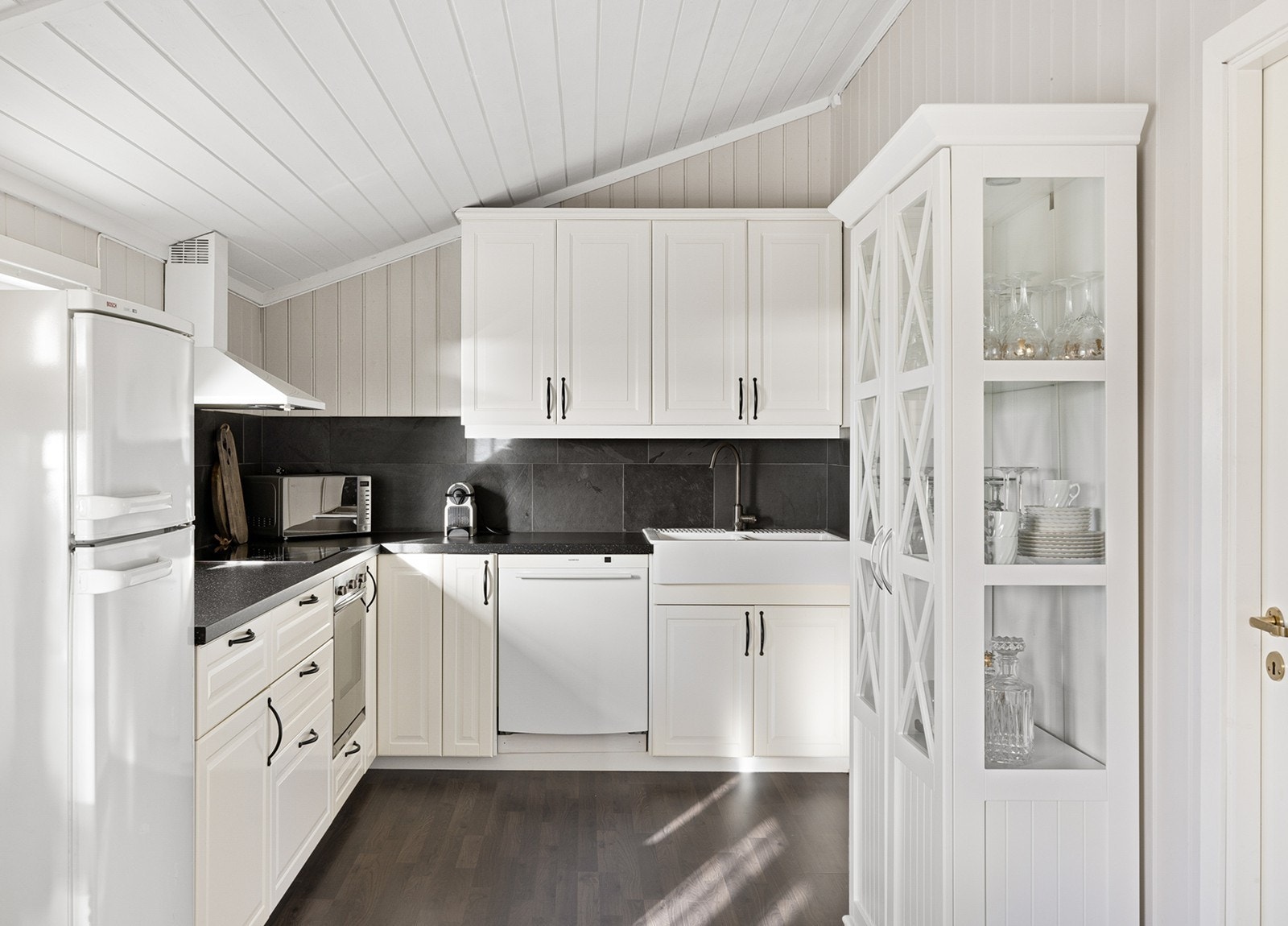 Koselig kjøkkeninnredning med hvite fronter og laminert benkeplate med nedfelt oppvaskkum med ett-greps blandebatteri. Opplegg for oppvaskmaskin, integrert komfyr og nedfelt platetopp, og det er tilkoblet en ventilator over kokeplass. Galleribilde