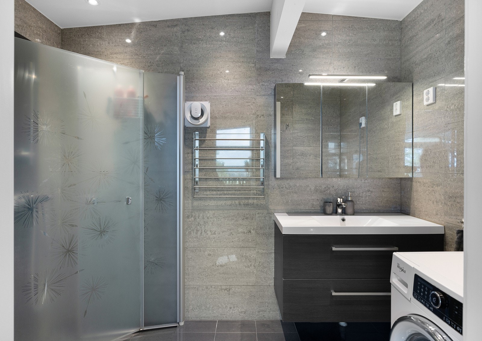 Flott flislagt bad med underliggende gulvvarme, opplegg for vaskemaskin og downlights i himling. Innredningen består av et underskap/overskap, servant, speil, lys og stikk. En dusjsone med innfellbare dusjdører. Galleribilde