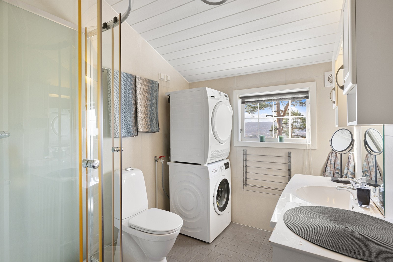 Et lyst flislagt bad med underliggende gulvvarme. Innredningen består av et underskap/overskap, servant, speil og armatur med stikk og et dusjkabinett. Opplegg for vaskemaskin og tørketrommel, og det er tilkoblet et vannklosett. Galleribilde