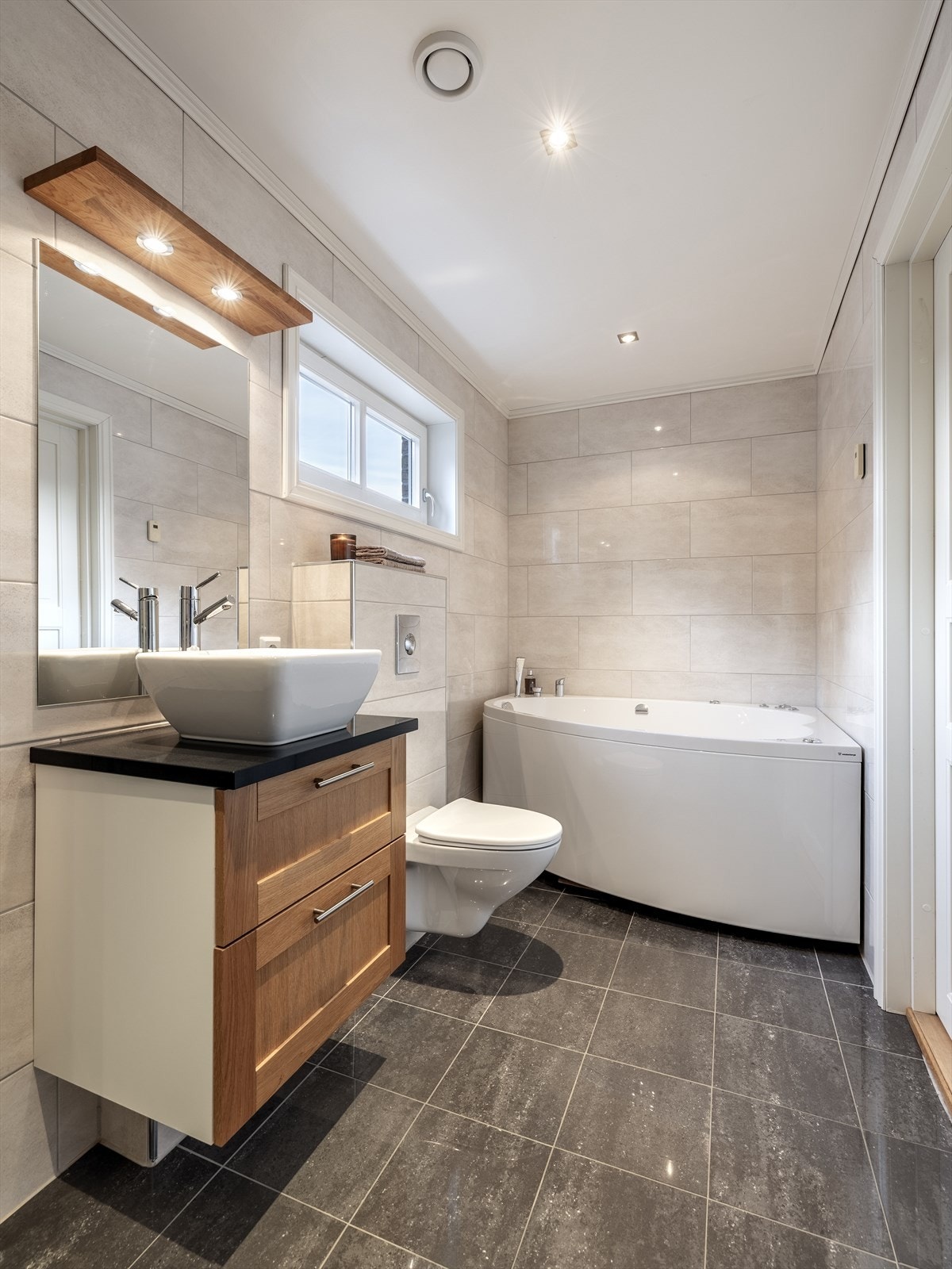 Badet inneholder dusjhjørne med innfellbare glassdører, badekar, veggmontert toalett og baderomsinnredning med servant, skuffer og speil. Galleribilde