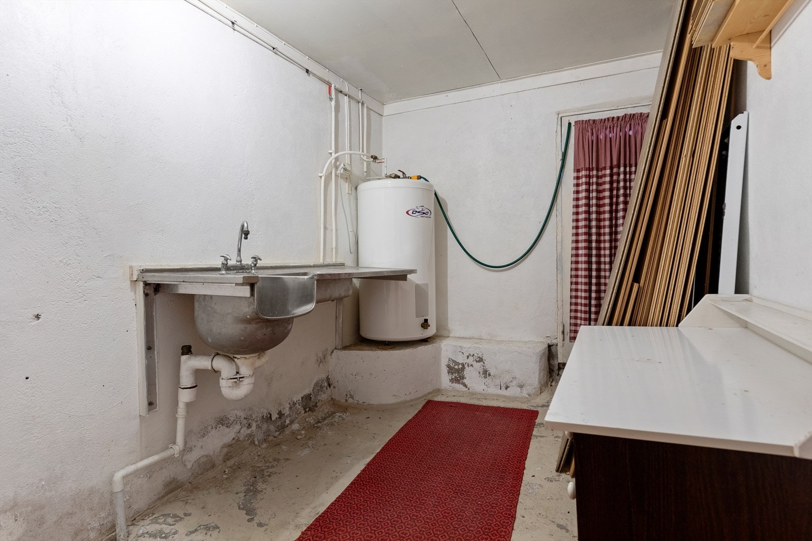 Vaskerom i kjelleretasjen har betonggulv og pussede murvegger | rommet er utstyrt med stålbenk med oppvaskkum, varmtvannsbereder og opplegg for vaskemaskin Galleribilde