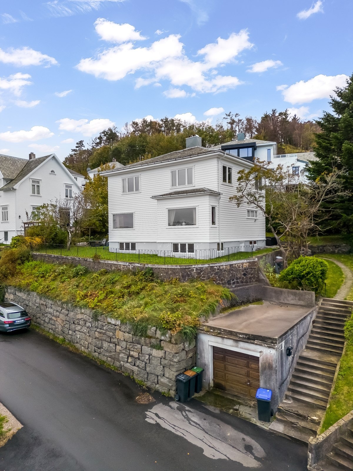 Enebolig bygd i funksjonalistisk byggestil - massiv steinmur mot veien og "Ålesundmur" ved huset. Garasje ved trappa som fører opp til boligen. Galleribilde