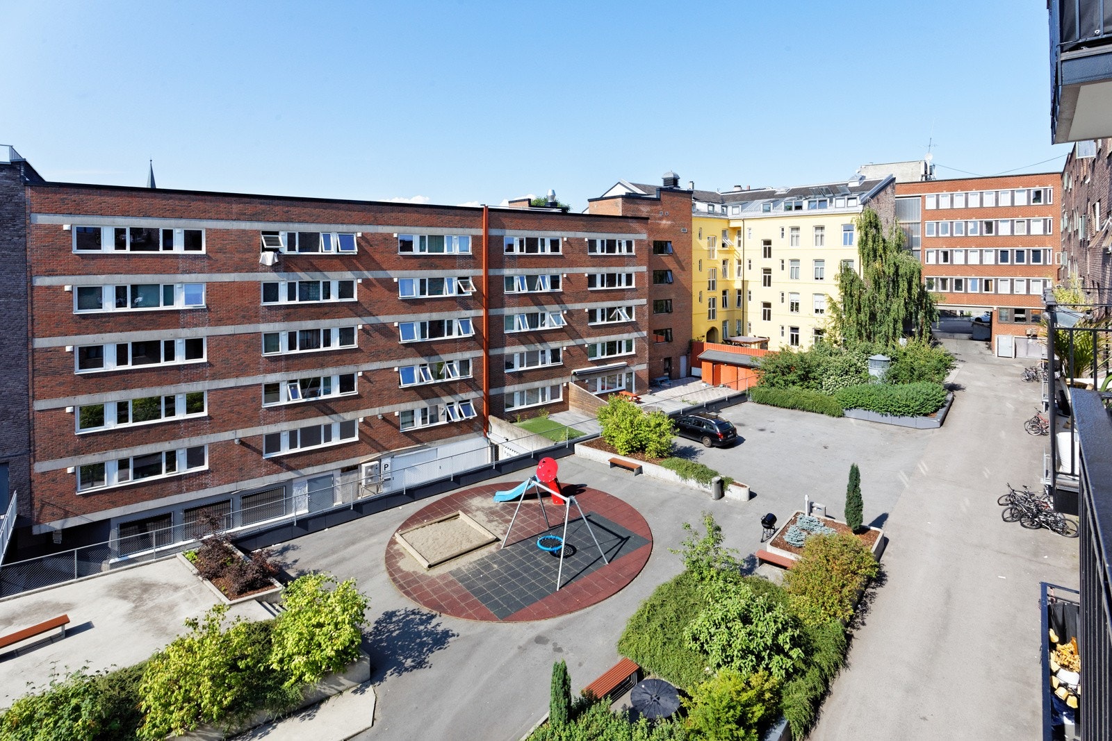 Pent opparbeidet bakgård med asfalterte områder, gressplener, sittegrupper, lekestativ, diverse busker og beplantinger. Galleribilde