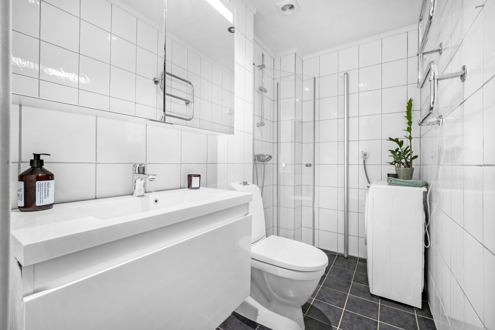 Rommet har grå fliser på gulv med gulvvarme, hvite fliser på veggene, downlights i taket og avtrekk fra elektrisk vifte. Innredningen består av en heldekkende servant på hvit skuffeseksjon, speilskap og elektrisk håndkletørker. Galleribilde