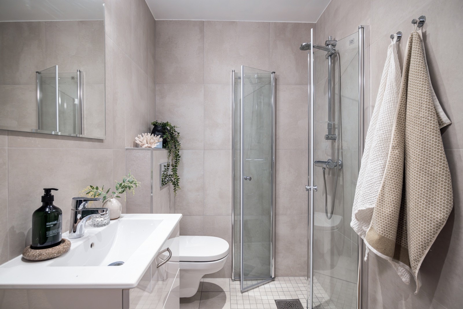 Moderne baderomsinnredning med skuffer og heldekkende servant, speil med belysning over. Baderommet er også utstyrt med dusjhjørne med innfellbare glassdører og vegghengt toalett. Galleribilde