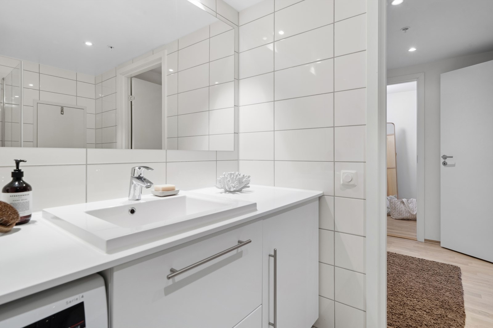 Badet er innredet med: Benkeplate med nedfelt servant. Underskap med glatte, folierte fronter. Speil på vegg over servant. Veggmontert klosett med utenpåliggende sisterne og opplegg for vaskemaskin. Galleribilde