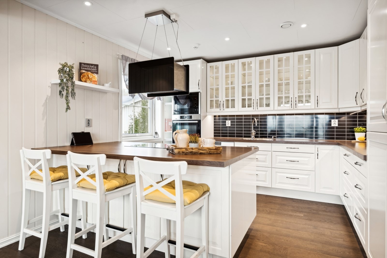 Innredningen er utført med hvite profilerte fronter, enkelte overskap med glassfelt, laminat benkeplate med underlimt vask og Kitchenboard på vegg over benkeplaten Galleribilde