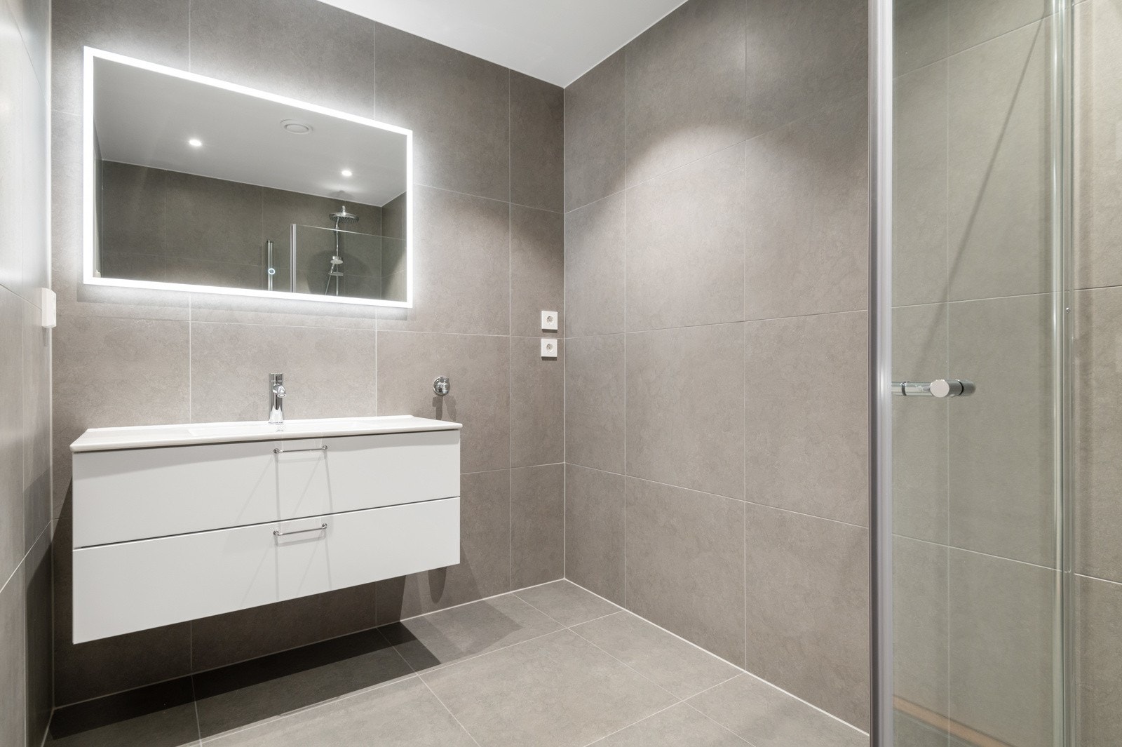 Flislagt bad utstyrt med - Servant m/lagring, speil m/lys, vegghengt toalett, dusjhjørne, downlights og opplegg for vaskemaskin Galleribilde