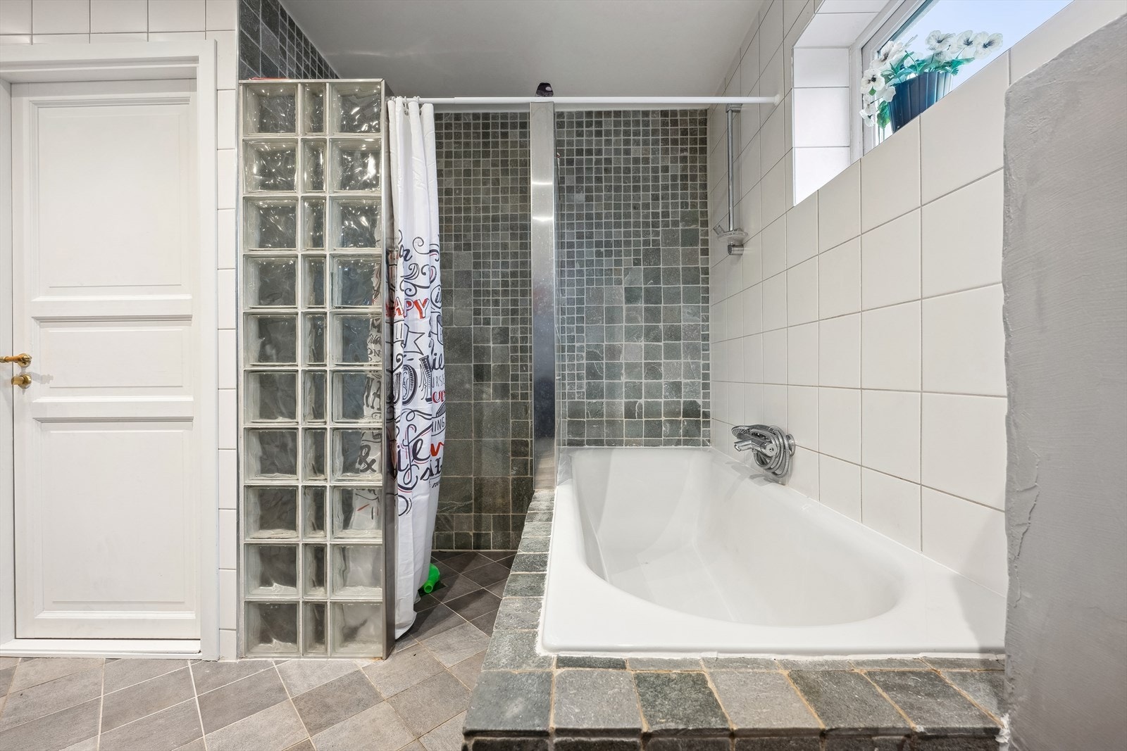 Helfliset baderom med nedfelt servant på skapinnredning med hvite fronter, overskap, speil med lys, veggmontert toalett, dusjvegger/hjørne samt et innmurt badekar. Galleribilde